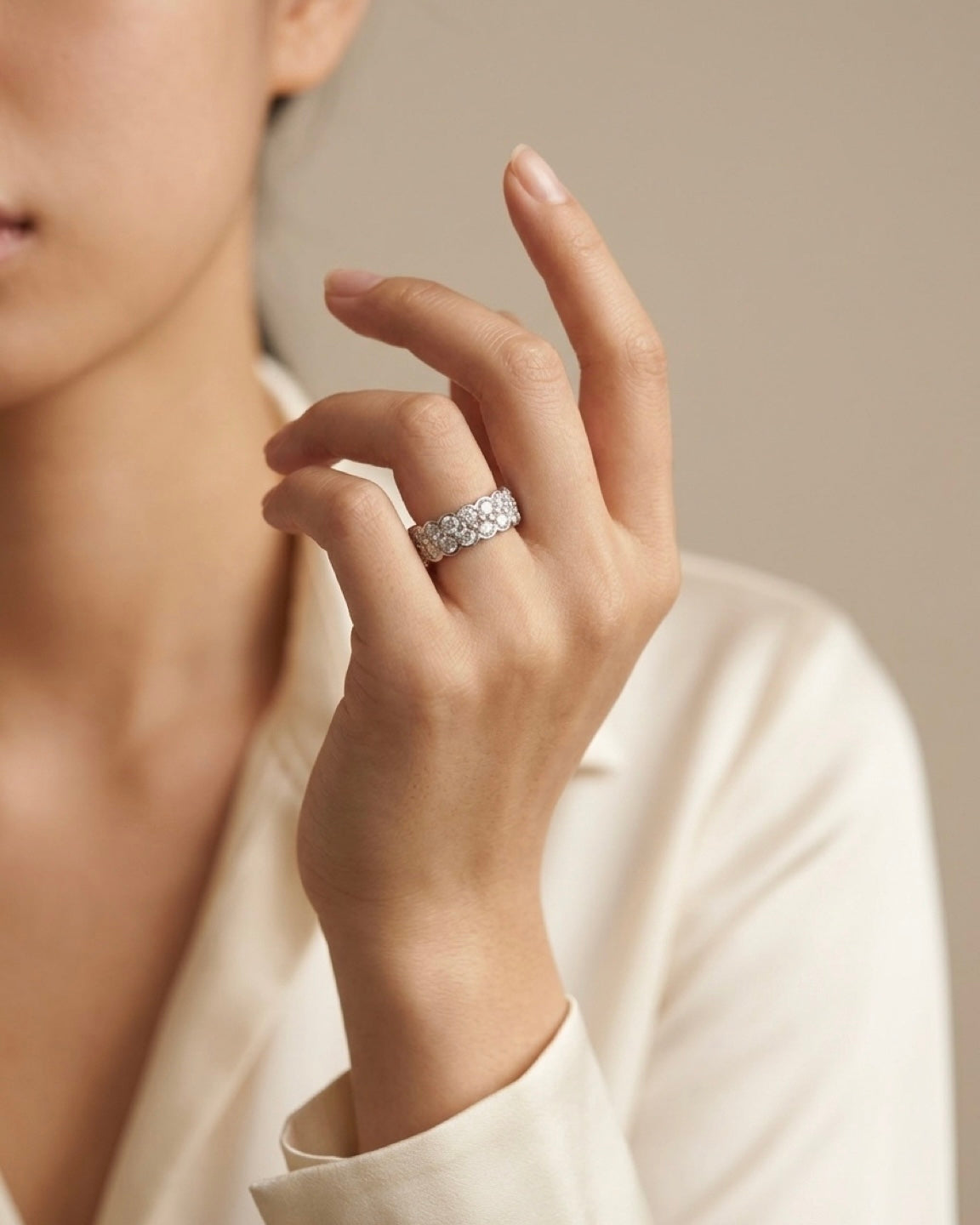 A close-up of a woman's hand wearing a silver band ring set with small clear stones, against a soft beige background.